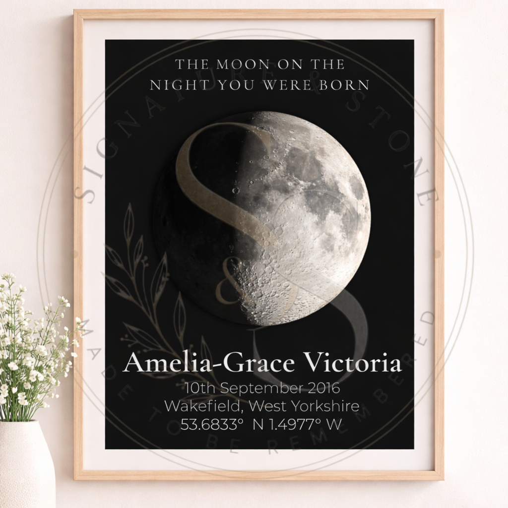 A framed personalized moon phase birth poster displayed on a light wall next to a small potted plant. The poster features a black background with a detailed grayscale moon illustration, white serif text reading 'THE MOON ON THE NIGHT YOU WERE BORN' and 'Amelia-Grace Victoria' with birth details below, and a light wood frame.