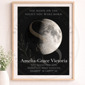A framed personalized moon phase birth poster displayed on a light wall next to a small potted plant. The poster features a black background with a detailed grayscale moon illustration, white serif text reading 'THE MOON ON THE NIGHT YOU WERE BORN' and 'Amelia-Grace Victoria' with birth details below, and a light wood frame.